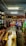 A small shop or market interior bustling with activity, featuring several people interacting at the counter. The shelves are stocked with a variety of goods, including bottles and packaged items. Overhead lighting brightens the space, and a ceiling fan can be seen.