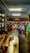 A small shop or market interior bustling with activity, featuring several people interacting at the counter. The shelves are stocked with a variety of goods, including bottles and packaged items. Overhead lighting brightens the space, and a ceiling fan can be seen.