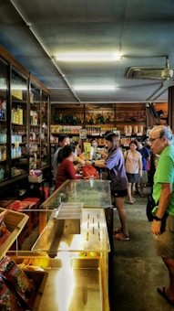 A small shop or market interior bustling with activity, featuring several people interacting at the counter. The shelves are stocked with a variety of goods, including bottles and packaged items. Overhead lighting brightens the space, and a ceiling fan can be seen.