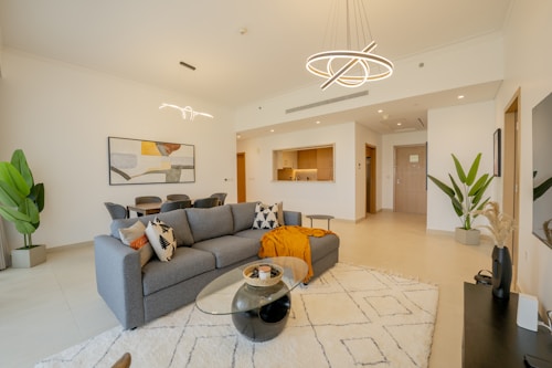 A modern living room featuring a grey sectional sofa adorned with decorative cushions and a mustard throw blanket. A glass coffee table is positioned in front of the sofa, placed on a large geometric-patterned rug. To the left, there's a dining area with a wooden table and black chairs. The room is decorated with potted plants and abstract wall art. Elegant light fixtures with unique designs hang from the ceiling, adding a contemporary touch. The overall space is open and bright, with neutral tones and minimalistic decor.