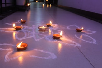 Several small clay oil lamps are lit and placed on the floor, surrounded by intricate white patterns. The scene is set indoors with a reflective surface and additional lamps visible in the background, creating a warm, festive atmosphere.
