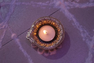 An intricately designed brass or metal holder contains a lit candle, casting a warm glow. The surface beneath features subtle decorative patterns in soft, muted tones.