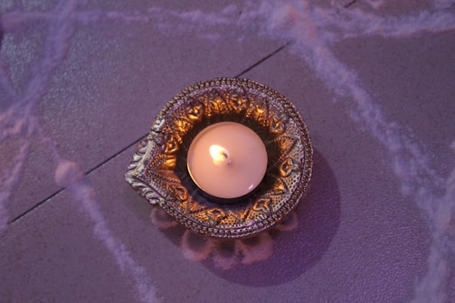 An intricately designed brass or metal holder contains a lit candle, casting a warm glow. The surface beneath features subtle decorative patterns in soft, muted tones.