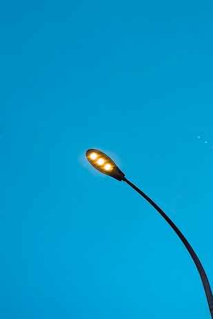 A streetlight with glowing lamps against a clear blue sky, featuring a sleek, modern design with a curved pole extending upwards.