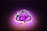 A stylized neon sign shaped like a graphical depiction of Lord Ganesha, with swirling lines and artistic curves. The sign emits a warm glow, combining shades of pink and purple, against a dark background.