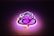A stylized neon sign shaped like a graphical depiction of Lord Ganesha, with swirling lines and artistic curves. The sign emits a warm glow, combining shades of pink and purple, against a dark background.