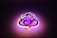 A stylized neon sign shaped like a graphical depiction of Lord Ganesha, with swirling lines and artistic curves. The sign emits a warm glow, combining shades of pink and purple, against a dark background.