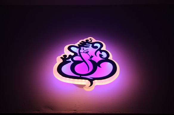 A stylized neon sign shaped like a graphical depiction of Lord Ganesha, with swirling lines and artistic curves. The sign emits a warm glow, combining shades of pink and purple, against a dark background.