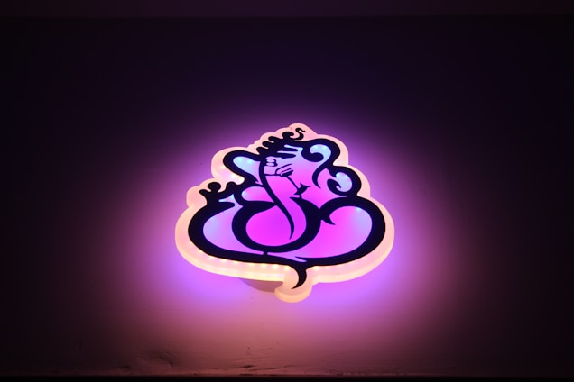 A stylized neon sign shaped like a graphical depiction of Lord Ganesha, with swirling lines and artistic curves. The sign emits a warm glow, combining shades of pink and purple, against a dark background.