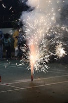 Fireworks are being set off on a pavement, creating bright sparks and smoke. People are gathered around, watching the display, with some individuals visible in the background.