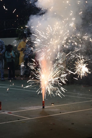 Fireworks are being set off on a pavement, creating bright sparks and smoke. People are gathered around, watching the display, with some individuals visible in the background.