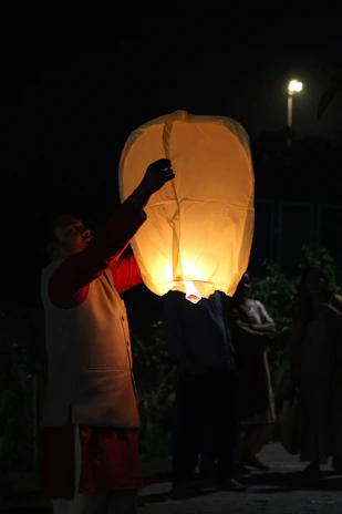 A smiling person holding a glowing lantern during an evening spiritual gathering outdoors.