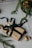 A gift wrapped in brown paper is adorned with a dark green ribbon and decorative golden accents. The package is set on a light-colored fabric surface, with pine branches scattered around, suggesting a festive or holiday theme. A shiny heart ornament rests nearby.