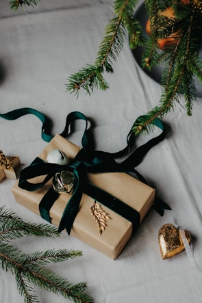 A gift wrapped in brown paper is adorned with a dark green ribbon and decorative golden accents. The package is set on a light-colored fabric surface, with pine branches scattered around, suggesting a festive or holiday theme. A shiny heart ornament rests nearby.