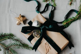 A gift wrapped in brown paper and adorned with a dark green ribbon, forest greenery, and small decorative bells. The scene includes evergreen branches and a golden star ornament, all placed on a light-colored surface.
