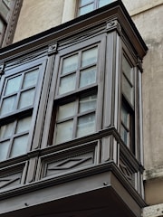 A detailed view of a wooden or painted bay window with decorative trim and multiple glass panes. The window is set against a smooth, light-colored wall with signs of weathering.
