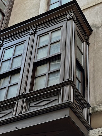 A detailed view of a wooden or painted bay window with decorative trim and multiple glass panes. The window is set against a smooth, light-colored wall with signs of weathering.