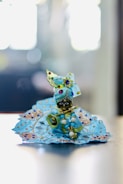 A small, intricately decorated idol dressed in vibrant blue clothing, adorned with beads and embroidery, is seated. The figure is holding a golden flute and is embellished with colorful jewels. The background is softly blurred, giving focus to the detailed craftsmanship of the idol.