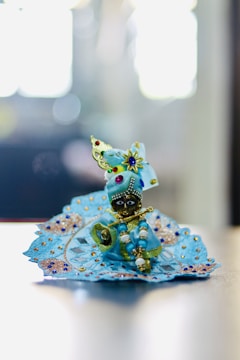 A small, intricately decorated idol dressed in vibrant blue clothing, adorned with beads and embroidery, is seated. The figure is holding a golden flute and is embellished with colorful jewels. The background is softly blurred, giving focus to the detailed craftsmanship of the idol.