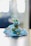 A small, intricately decorated idol dressed in vibrant blue clothing, adorned with beads and embroidery, is seated. The figure is holding a golden flute and is embellished with colorful jewels. The background is softly blurred, giving focus to the detailed craftsmanship of the idol.