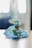 A small, intricately decorated idol dressed in vibrant blue clothing, adorned with beads and embroidery, is seated. The figure is holding a golden flute and is embellished with colorful jewels. The background is softly blurred, giving focus to the detailed craftsmanship of the idol.