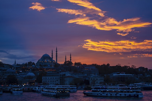 A stunning sunset casts vibrant orange and purple hues across the sky, silhouetting the iconic skyline with a prominent mosque and its minarets. Boats are gently floating on the water, contributing to the serene and picturesque scene.