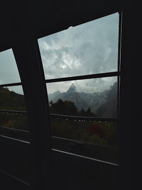 View of a mountain range with snow-covered peaks seen through a large window. The scene includes a cloudy sky with varying shades of gray, adding a dramatic flair. Trees in the foreground provide a natural border, and the contrast between the dark interior and bright exterior adds depth.