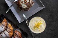 A flat lay of various desserts. On the top left, a plate holds a decadent chocolate-covered dessert with nuts on top, accompanied by a fork. Below it, there is a black rectangular plate with sliced croissants dusted with powdered sugar. On the right side, a glass contains a creamy dessert topped with lemon zest.