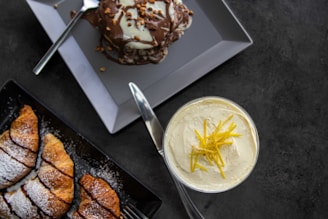 A flat lay of various desserts. On the top left, a plate holds a decadent chocolate-covered dessert with nuts on top, accompanied by a fork. Below it, there is a black rectangular plate with sliced croissants dusted with powdered sugar. On the right side, a glass contains a creamy dessert topped with lemon zest.