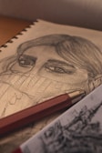 A detailed pencil sketch of a person's face partially concealed by a sweater collar is displayed on a notepad. The focus is on the eyes and hair, with intricate shading and texture. A red mechanical pencil lies across the pad, adding a splash of color to the monochrome artwork. Another piece of art is partially visible in the foreground.