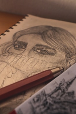 A detailed pencil sketch of a person's face partially concealed by a sweater collar is displayed on a notepad. The focus is on the eyes and hair, with intricate shading and texture. A red mechanical pencil lies across the pad, adding a splash of color to the monochrome artwork. Another piece of art is partially visible in the foreground.