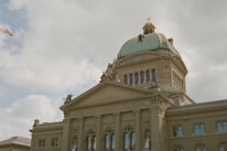 A grand architectural building featuring classic design elements such as columns, arched windows, and a large dome with a cross on top. Statues are positioned on the rooftop, adding to the ornate style. The inscription 'Curia Confoederationis Helveticae' is visible above the columns. The sky is overcast with thick clouds, and a crane is seen in the background.