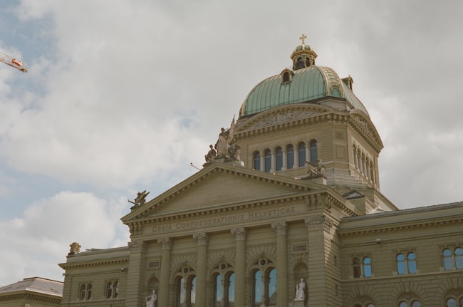 A grand architectural building featuring classic design elements such as columns, arched windows, and a large dome with a cross on top. Statues are positioned on the rooftop, adding to the ornate style. The inscription 'Curia Confoederationis Helveticae' is visible above the columns. The sky is overcast with thick clouds, and a crane is seen in the background.