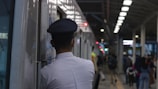 A train station platform with a person in uniform, possibly a conductor or security, standing next to a train. The platform is busy with people walking in various directions. Overhead lighting and structural elements of the station are visible.