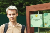 A young person with light hair is standing outdoors, wearing a light brown shirt and a backpack. Next to them is a wooden structure with green panels featuring decorative designs, including leaves and text in Russian.