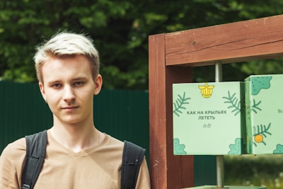 A young person with light hair is standing outdoors, wearing a light brown shirt and a backpack. Next to them is a wooden structure with green panels featuring decorative designs, including leaves and text in Russian.