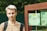 A young person with light hair is standing outdoors, wearing a light brown shirt and a backpack. Next to them is a wooden structure with green panels featuring decorative designs, including leaves and text in Russian.