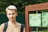 A young person with light hair is standing outdoors, wearing a light brown shirt and a backpack. Next to them is a wooden structure with green panels featuring decorative designs, including leaves and text in Russian.
