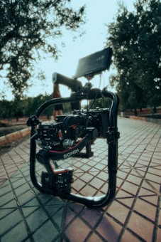 A professional camera stabilizer setup is positioned on a tiled pathway, surrounded by greenery. The setup includes a RONIN gimbal with a mounted camera, and a monitor attached at the top. The background features blurred vegetation, indicating an outdoor environment with natural lighting.