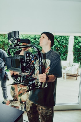 A person wearing a black beanie and printed T-shirt operates a professional camera mounted on a stabilizer. The setting appears to be indoors with natural light streaming in, and greenery visible through large windows. Other individuals are partially visible in the background.