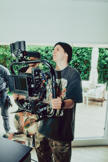A person wearing a black beanie and printed T-shirt operates a professional camera mounted on a stabilizer. The setting appears to be indoors with natural light streaming in, and greenery visible through large windows. Other individuals are partially visible in the background.