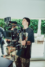 A person wearing a black beanie and printed T-shirt operates a professional camera mounted on a stabilizer. The setting appears to be indoors with natural light streaming in, and greenery visible through large windows. Other individuals are partially visible in the background.
