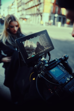 A professional video camera setup captures a scene on a city street. In the background, a woman stands out of focus while the camera monitor displays her clearly framed against urban buildings. The equipment appears to be high-end, suggesting a film or photography production.