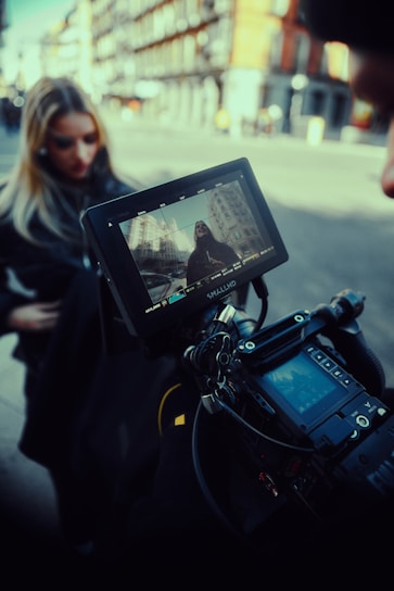A professional video camera setup captures a scene on a city street. In the background, a woman stands out of focus while the camera monitor displays her clearly framed against urban buildings. The equipment appears to be high-end, suggesting a film or photography production.