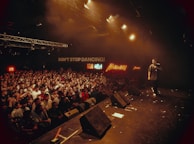 A lively concert venue filled with an enthusiastic crowd facing the stage. Bright lights illuminate the scene, casting a warm glow over the audience. A performer holds a microphone, engaging with the crowd. Neon signs and a message reading 'DON'T STOP DANCING!' add to the vibrant atmosphere.
