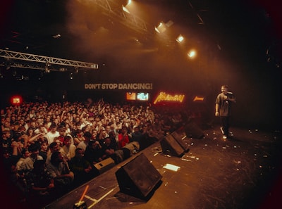 A lively concert venue filled with an enthusiastic crowd facing the stage. Bright lights illuminate the scene, casting a warm glow over the audience. A performer holds a microphone, engaging with the crowd. Neon signs and a message reading 'DON'T STOP DANCING!' add to the vibrant atmosphere.