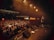 A lively concert venue filled with an enthusiastic crowd facing the stage. Bright lights illuminate the scene, casting a warm glow over the audience. A performer holds a microphone, engaging with the crowd. Neon signs and a message reading 'DON'T STOP DANCING!' add to the vibrant atmosphere.