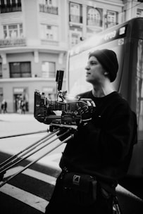 A person dressed in dark clothing is holding a camera rig with multiple stabilizing supports. The background features urban architecture with multiple windows and a nearby vehicle. The photo is in black and white, giving it a classic and artistic feel.