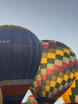 Several hot air balloons are in the process of being inflated. They are colorful and have different patterns, including one with a checkered pattern in yellow, red, green, and black, and another with alternating blue and grey vertical stripes.