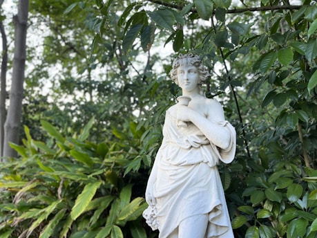 A classical white marble statue of a woman, adorned with curly hair and draped in a flowing robe, stands gracefully against a lush green backdrop of leaves and trees. The statue appears to be holding an object close to her chest.